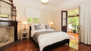 Un dormitorio con piso de madera, una cama grande con ropa blanca, dos mesitas de noche con lámparas, un espejo y vista al exterior a través de una puerta de cristal.