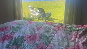 A bed with a floral bedspread, a window with curtains, and a view of a grassy area with two chairs and a body of water. - Relax & Unwind: Private Beachfront Escape with Stunning Views, Amazing! AC 1328 (Anahola)