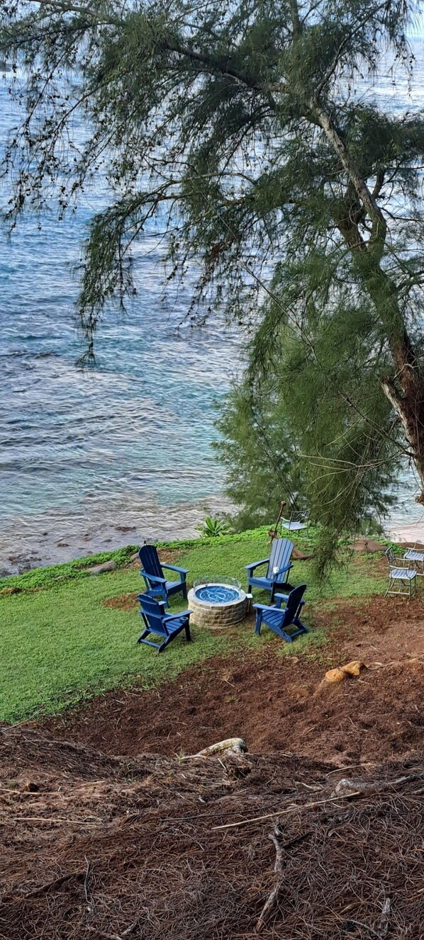 A lakeside setting with two blue Adirondack chairs and a small circular table, surrounded by a grassy area and a tree.