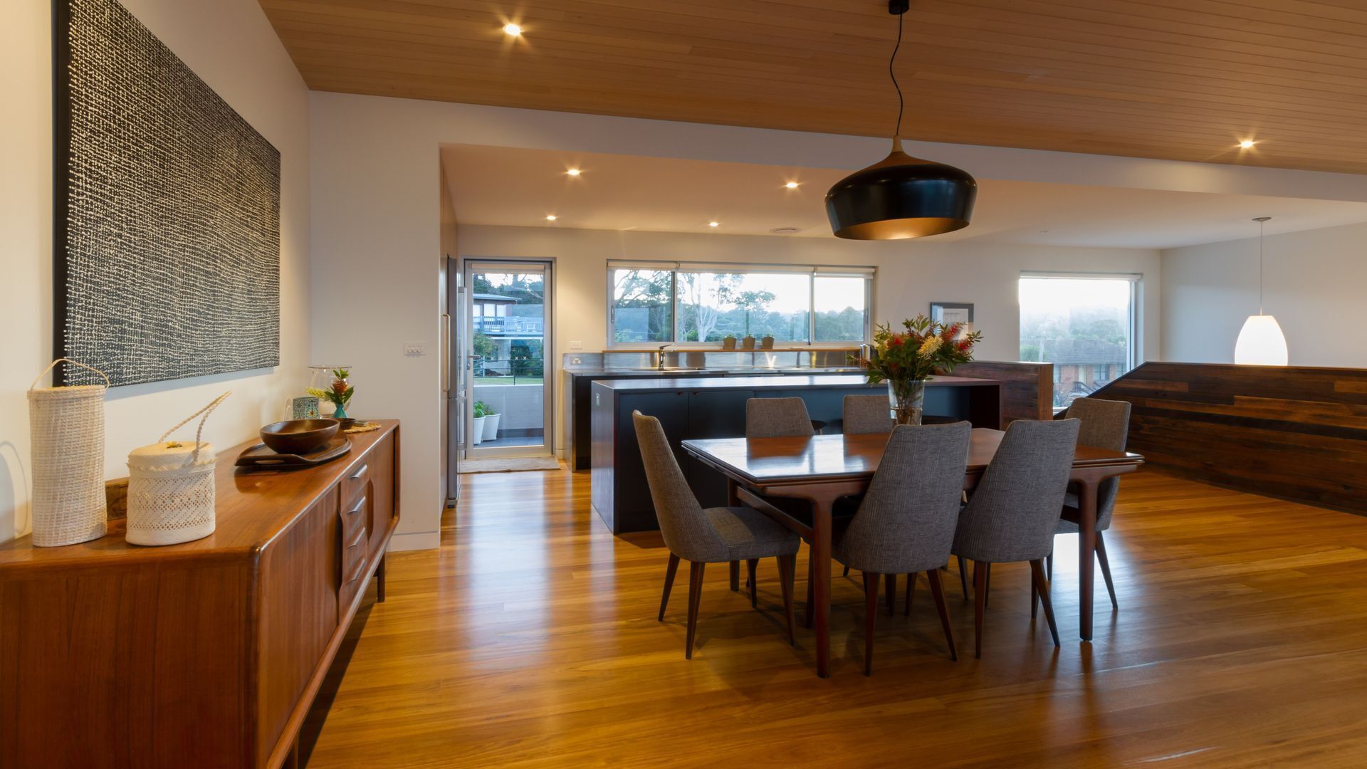 A modern kitchen with a dining area, wooden flooring, and a large window providing a view of the outdoors.
