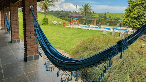 A hammock under a wooden pergola overlooking a pool area with lounge chairs and a thatched-roof structure.