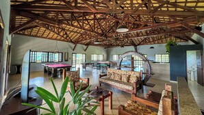 A spacious indoor area with a high ceiling, wooden beams, and large windows. There are multiple tables and a sofa with floral upholstery.