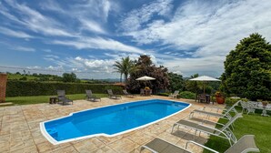 A rectangular swimming pool with a tiled deck, surrounded by lounge chairs and umbrellas, set against a backdrop of lush greenery and a clear sky.