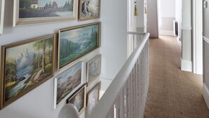 A hallway with a white railing, a herringbone-patterned carpet, and framed landscape paintings on the walls. - Stylish Luxury Let In Rye. Walk To Shops/Pubs/Restaurants + Near Beach (Rye)