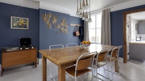 A dining area with a wooden table and chairs, a small side table with a bowl of fruit, and a wall-mounted decorative piece. - Stylish Luxury Let In Rye. Walk To Shops/Pubs/Restaurants + Near Beach (Rye)