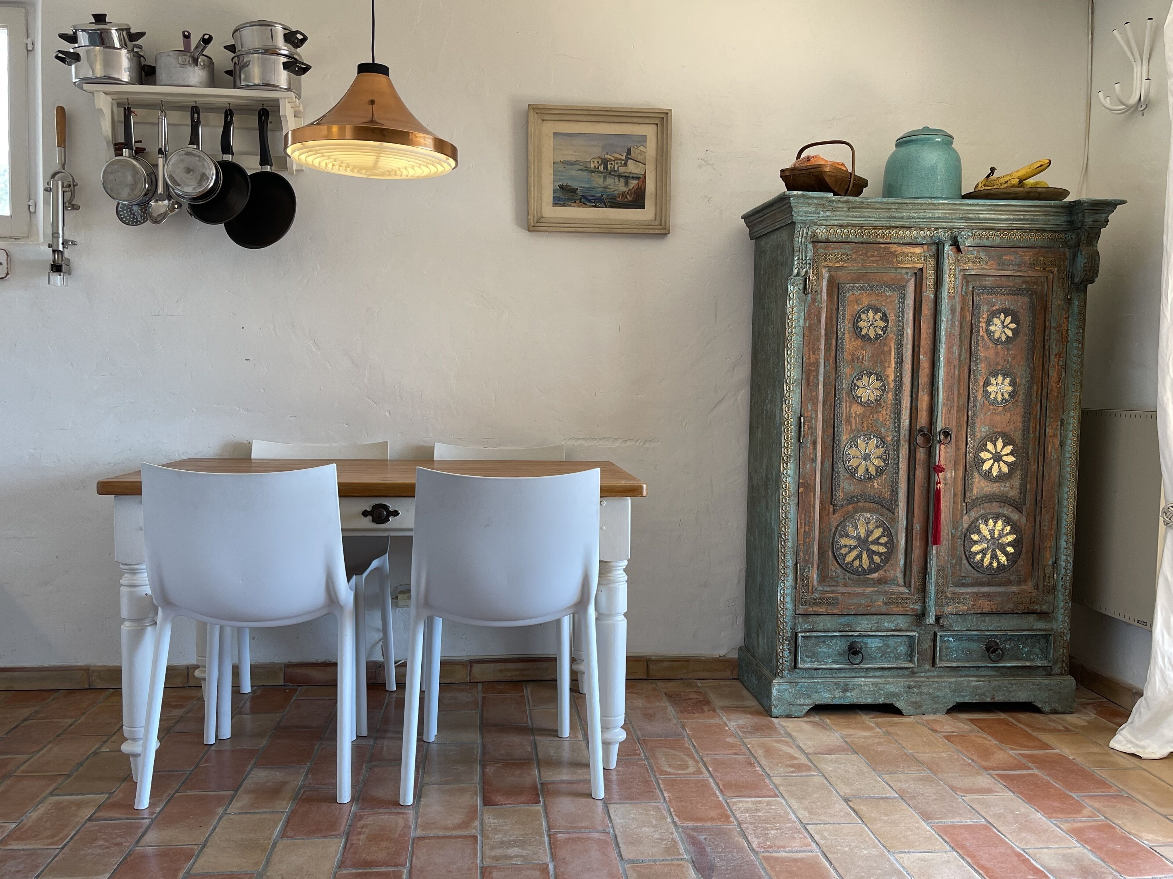 Een eetgedeelte met een houten tafel, witte stoelen, een hanglamp, een aan de muur bevestigde kruidenrek en een decoratieve kast met bloemenmotieven.