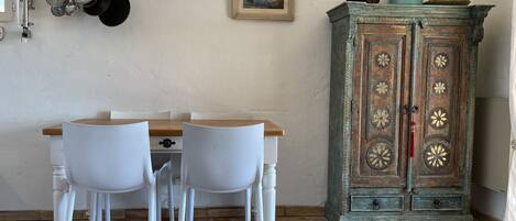 Een eetgedeelte met een houten tafel, witte stoelen, een hanglamp, een aan de muur bevestigde kruidenrek en een decoratieve kast met bloemenmotieven.