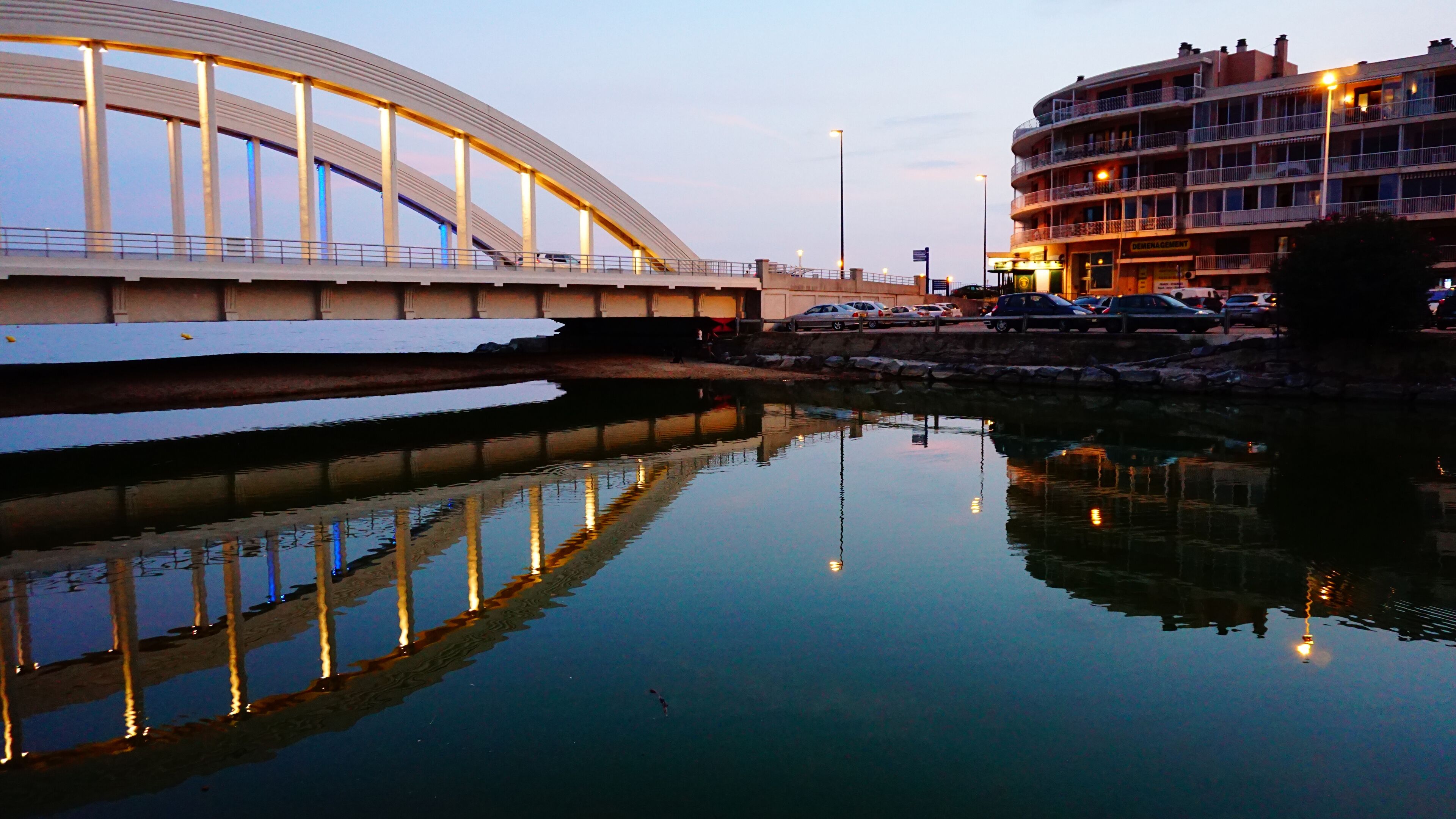 Un pont enjambant une étendue d’eau calme, dont le reflet ainsi que celui des bâtiments environnants se mirent dans l’eau.