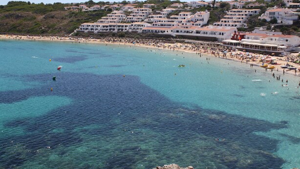Une ville côtière dotée d’une plage, d’une eau d’un bleu limpide et de bâtiments construits à flanc de colline.