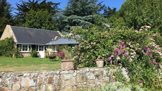 A stone house with a sloped roof, surrounded by lush greenery and flowering plants.