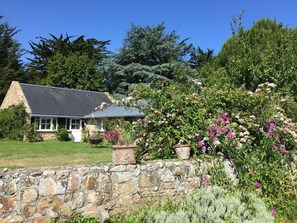 Una casa de piedra con techo inclinado, rodeada de abundante vegetación y plantas con flores.