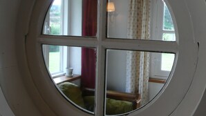 A room with a large circular window divided into four panes, a lamp, and a view of a green couch and a window with curtains.