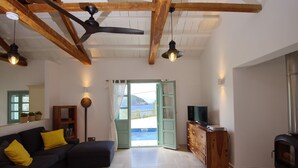 A living room with a dark sofa, wooden ceiling beams, a television on a wooden cabinet, and a view of the sea through an open door.