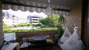 En balkon med et rundt bord, stole, en hængekøje og udsigt til en swimmingpool samt bygninger i baggrunden.