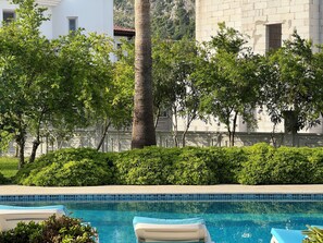 A swimming pool with lounge chairs, surrounded by lush greenery and a stone building in the background.