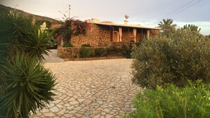 Un edificio de piedra con entrada de tejas, rodeado de vegetación y un muro de piedra.