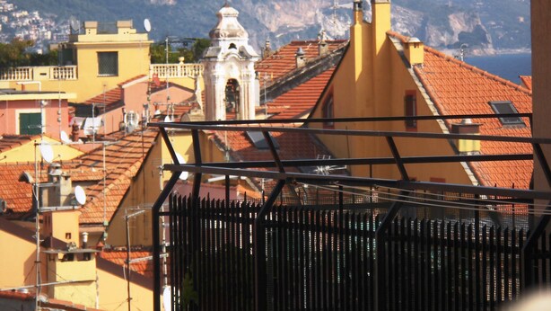 Un pueblo costero con edificios de tejas color terracota, una imponente torre de campanas y un telón de fondo montañoso.