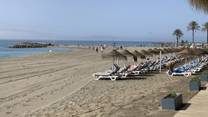 Una playa con filas de tumbonas y sombrillas de paja, un rompeolas rocoso y palmeras al fondo.
