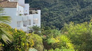 Un edificio blanco con varios balcones que dan a una exuberante ladera verde y montañas al fondo.