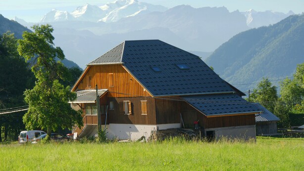 Una casa de madera con techo inclinado, rodeada de un campo cubierto de hierba y montañas al fondo.
