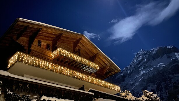 A traditional wooden chalet with a balcony adorned with lights, set against a snowy mountain backdrop. - Quiet, centrally located apartment with unobstructed views of the mountains + north face of the Eiger! (Grindelwald)