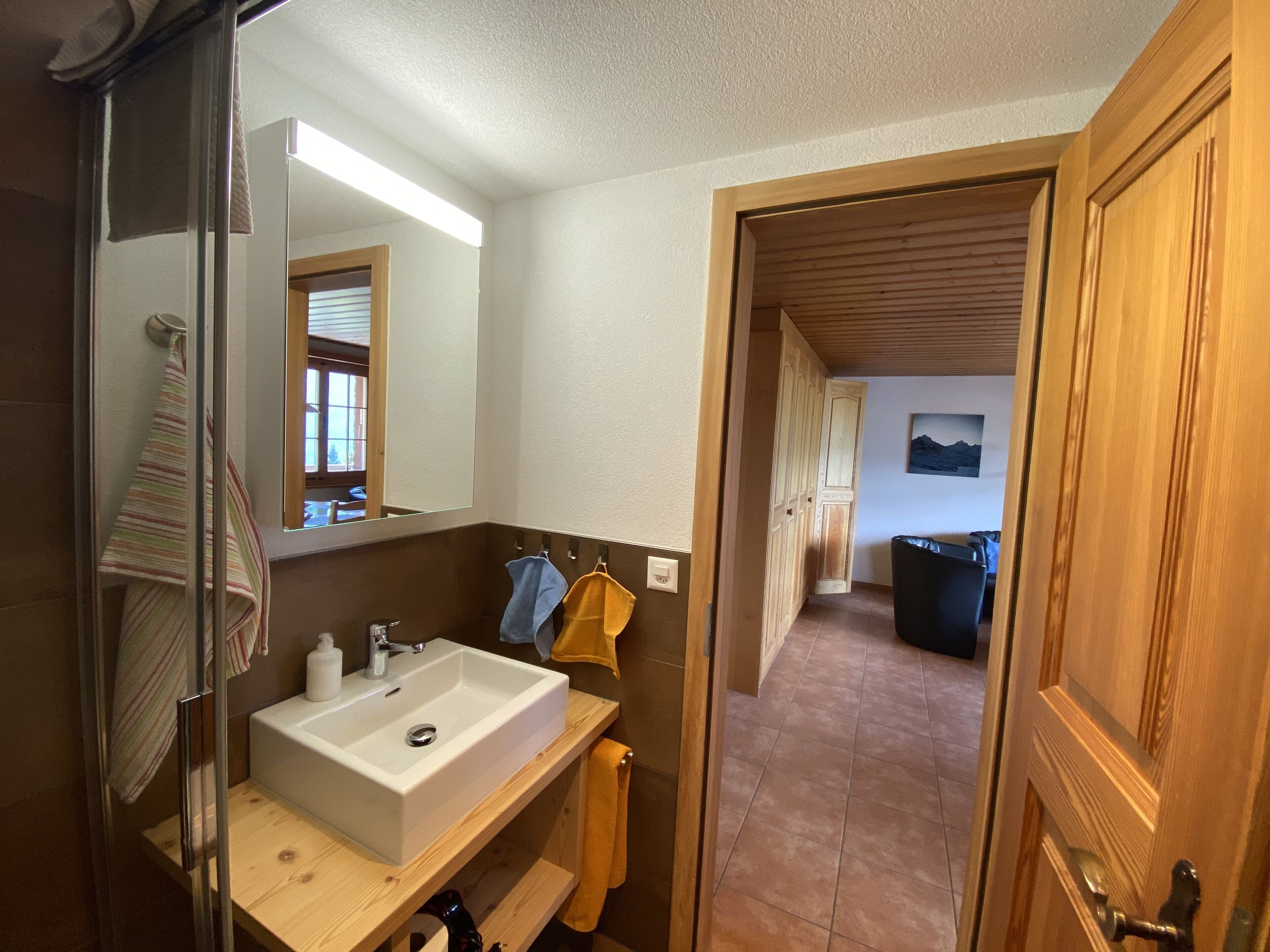 A bathroom with a white sink, wooden vanity, and a mirror. There is a towel rack with multiple towels and a wooden door leading to another room.
