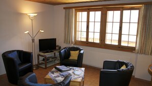 A living room with two black leather sofas, a wooden coffee table with magazines, a TV on a stand, and a window with curtains. - Quiet, centrally located apartment with unobstructed views of the mountains + north face of the Eiger! (Grindelwald)