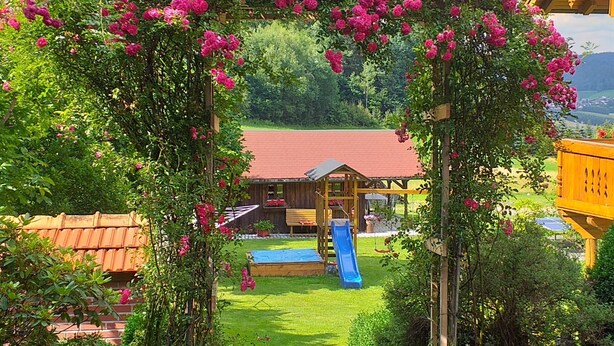 A wooden play structure with a slide and a small wooden building, surrounded by a garden with pink flowers and a stone pathway.