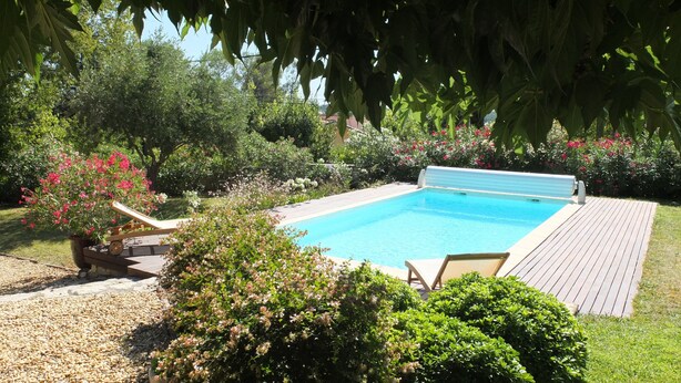 Une piscine rectangulaire entourée d’une terrasse en bois, située dans un jardin aménagé avec différents arbustes et plantes à fleurs.