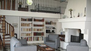 Un salon avec deux fauteuils gris, une table basse et une bibliothèque remplie de livres. On aperçoit un escalier au fond.
