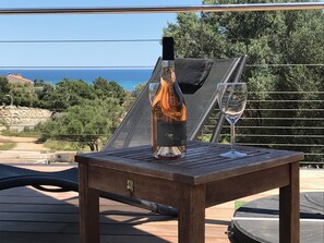 A bottle of wine and a glass on a wooden table with a view of the sea and greenery.