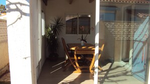 Eine sonnendurchflutete Terrasse mit einem Holztisch und -stühlen, einem Blumentopf und einem Fenster mit Blick ins Freie.