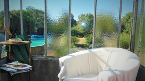 A room with a large window overlooking a pool and greenery. There is a chair and a table with magazines and a lamp.