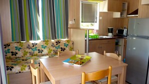 A compact kitchen with a wooden table, chairs, a sofa with a floral pattern, a refrigerator, and a window with curtains. - 2/4-person CLIMATIZED WOODEN CHALET CLOSE TO ALGAJOLA village views (Algajola)