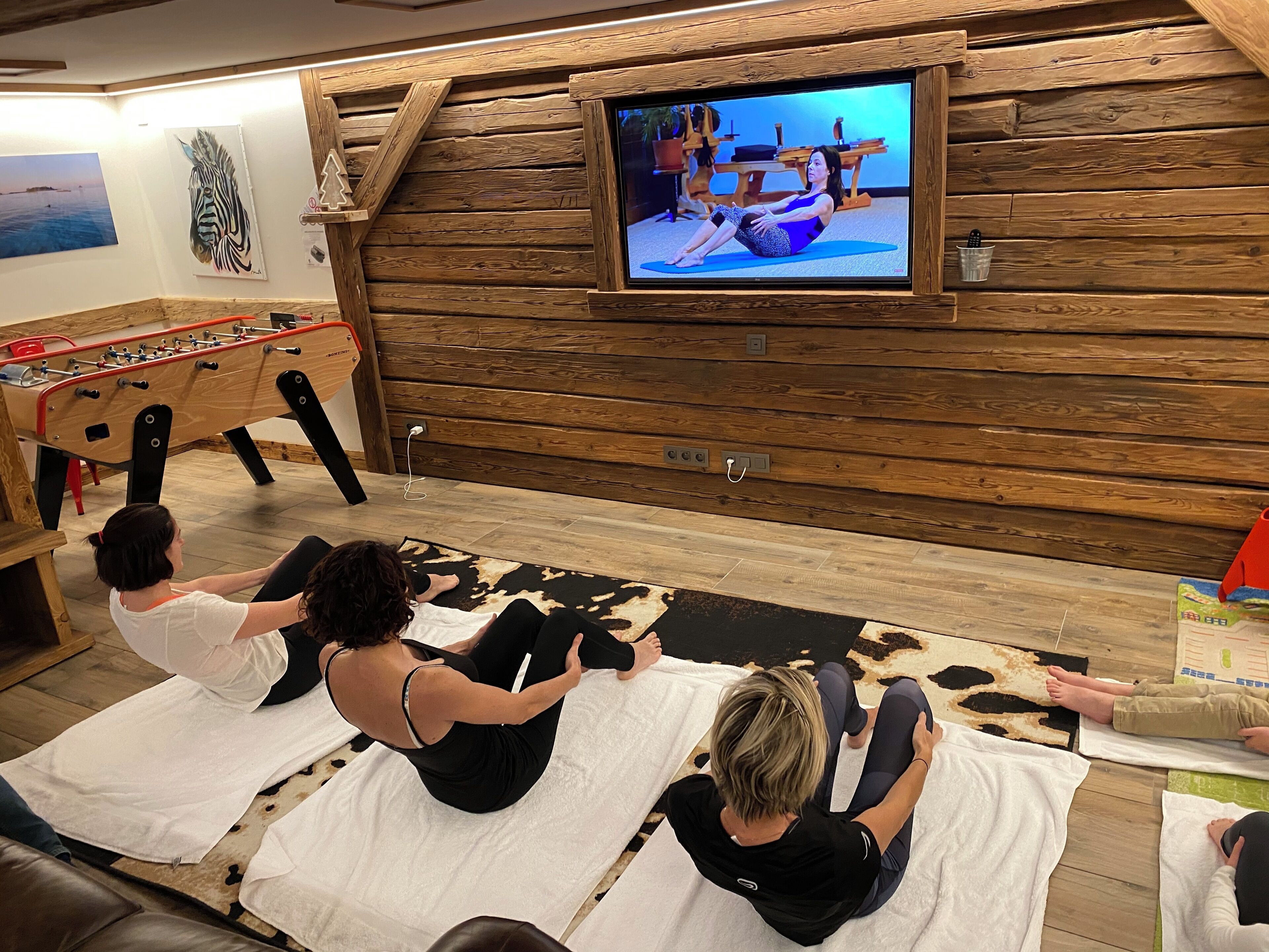 A room with wooden walls, a television displaying a yoga session, and a foosball table.