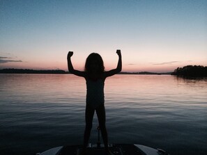 A child standing on a boat with arms raised, facing a calm lake at dusk.