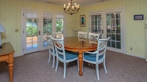 A dining room with a round wooden table, six white chairs with blue cushions, a chandelier, and double doors leading to a garden. - 4th Row Ocean Home w/ Private Pool (Hilton Head Island)