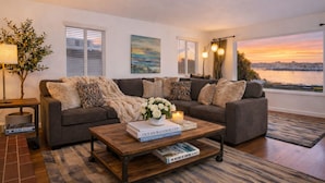 A living room with a sofa, coffee table, and books, a lamp, and a view of a sunset over water. - OCEANFRONT! BEST LOCATION! EXTRAORDINARY VIEWS! HOT TUB! FIRE PIT! (Santa Cruz)