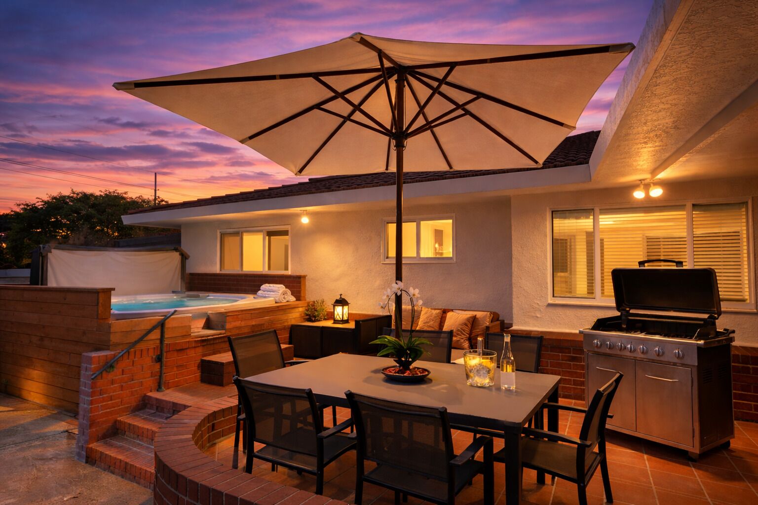 An outdoor patio with a table, chairs, a grill, a hot tub, and a large umbrella.