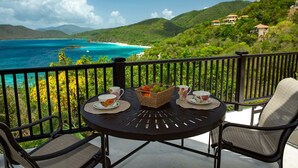 Un balcón con una mesa dispuesta para dos, con vista al mar turquesa y colinas verdes exuberantes.