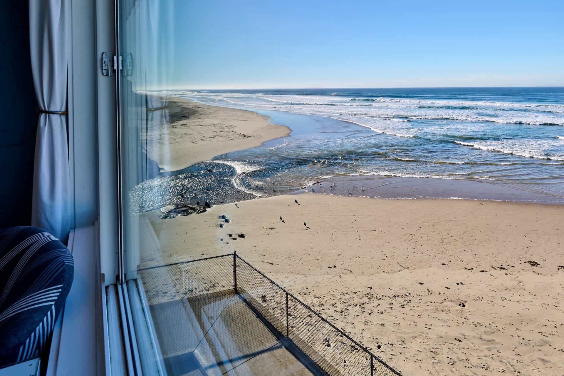 A beach view from a window with a metal railing, a clear sky, and the ocean.