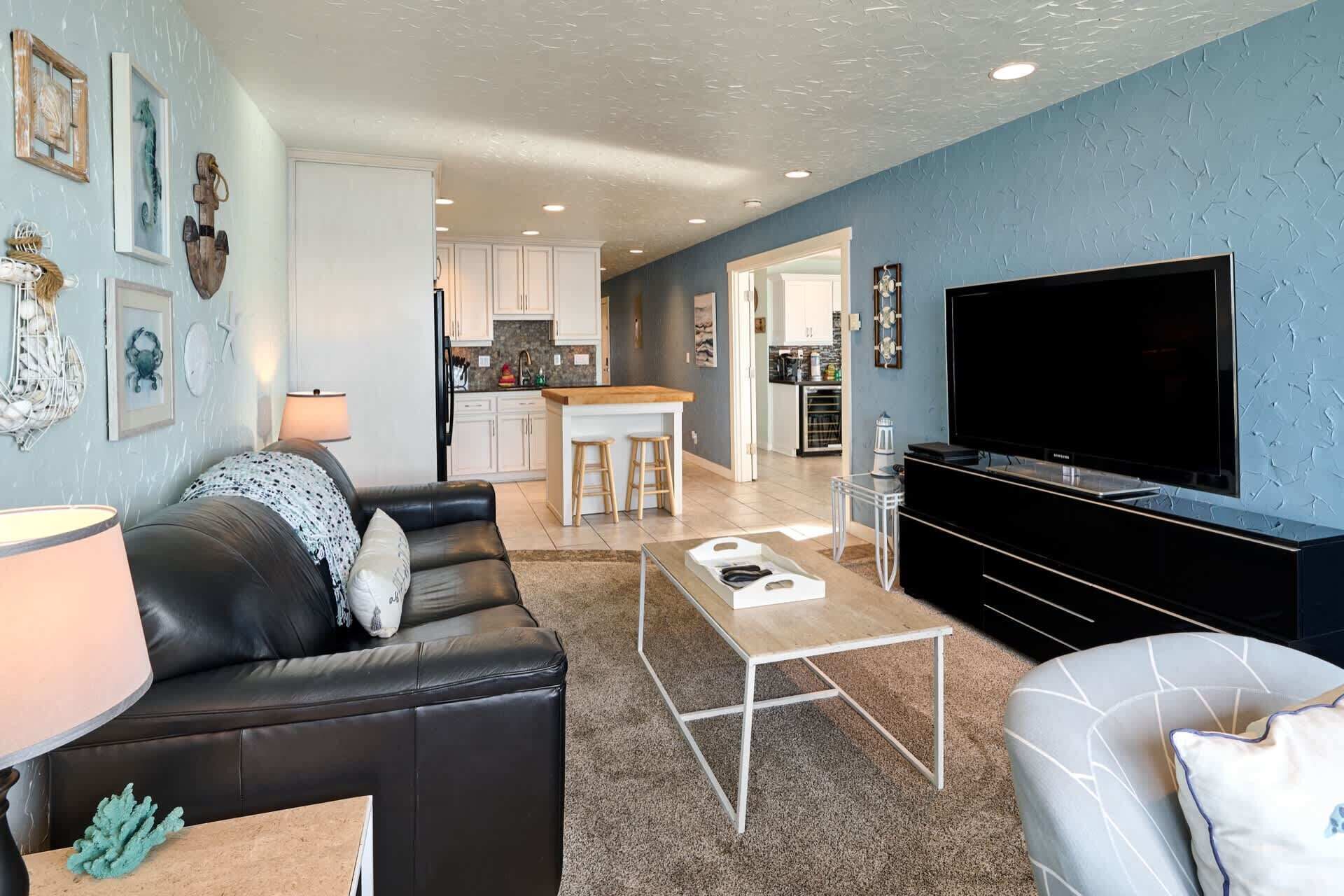 A living room with a black leather sofa, a coffee table, and a TV. There is a kitchen in the background with white cabinets and a dining area.