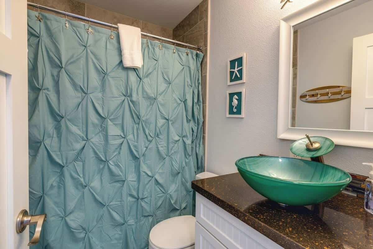 Un baño con cortina de ducha color turquesa, lavamanos de vidrio verde y un inodoro blanco.