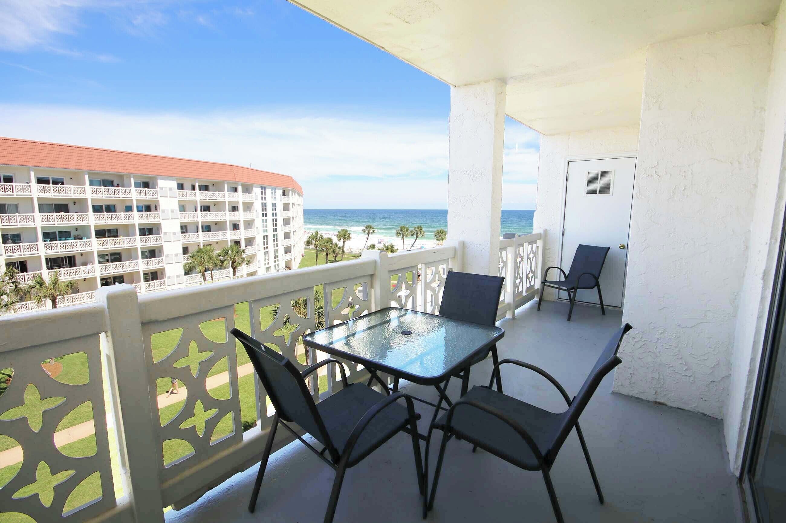 Uma varanda com mesa e cadeiras de vidro, com vista para uma praia e um edifício de hotel.