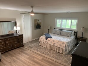 A bedroom with a bed, dresser, ceiling fan, window, and artwork on the wall.