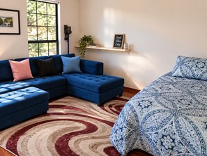 A modern living room with a sectional sofa, a patterned bed, a red and beige rug, and a shelf with a plant and a sign.
