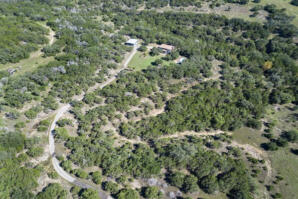 An aerial view of a property with a winding road, dense forest, and a house with a swimming pool.