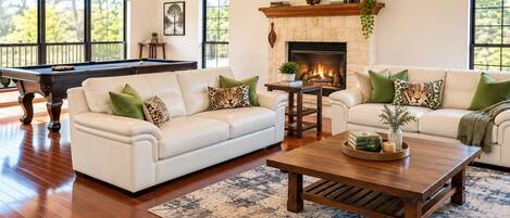 A spacious living room with a pool table, two sofas with decorative pillows, a fireplace, and a large window with a view of trees.