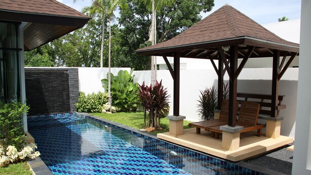 Ein Swimmingpool mit blauen Mosaikfliesen, umgeben von einem Garten mit verschiedenen Pflanzen und einem hölzernen Pavillon mit Sitzgelegenheiten.