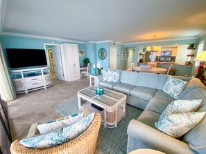 Sala de estar com sofá, mesa de centro e aparador de TV. Há uma área de jantar com mesa e cadeiras. O cômodo possui paredes azul claro e piso de azulejo.